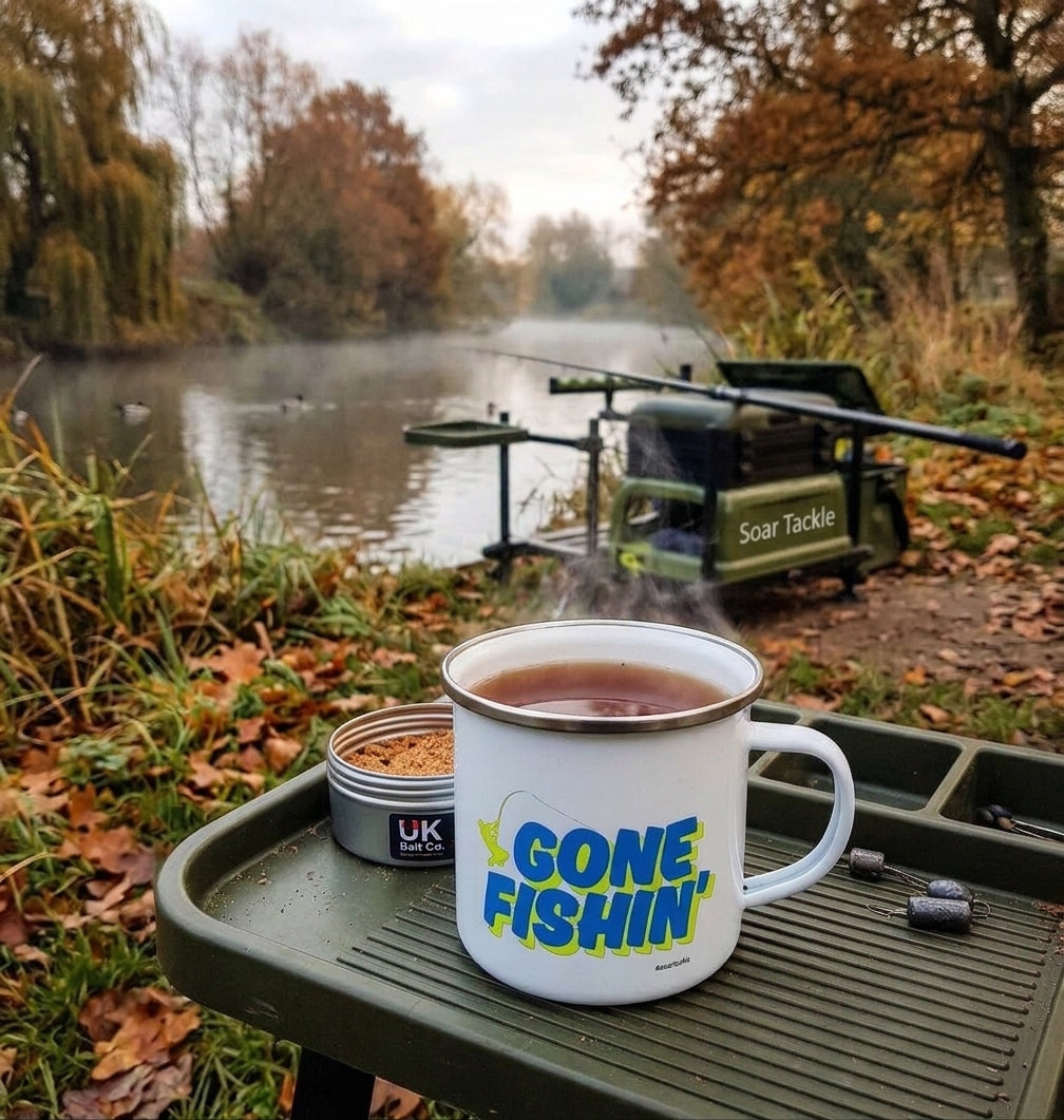 A white enamel camping mug with 'GONE FISHIN'' printed in blue and green sits on a green fishing table outdoors by a river, by Soar Tackle